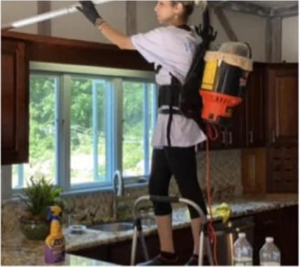 A Vcleaning cleaner on a step ladder with a backpack vacuum, dusting the top of kitchen cabinets